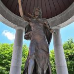 Bronze statue of a warrior holding a spear beneath a large, domed structure outdoors.