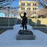 Bronze statue of a soldier stands on a paved walkway with a large building in the background.