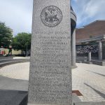 Granite memorial pillar with engraved names and seal, set outdoors under a blue sky.