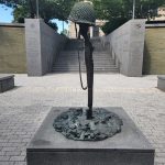 Abstract metal sculpture of a helmet stands on a stone base in an outdoor plaza.