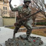 Bronze statue of a soldier holding a rifle, standing on rocks outdoors.