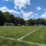 Green soccer field under a blue, cloudy sky with trees lining the background.